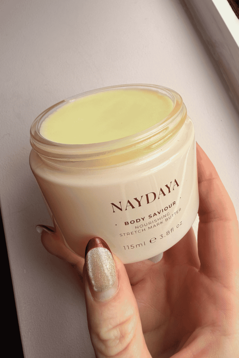 A hand with glittery silver nail polish holds a jar of NAYDAYA Body Saviour for stretch marks. The product is a creamy off-white color with a pale yellow cream visible at the top through a clear lid, set against a neutral light gray background.