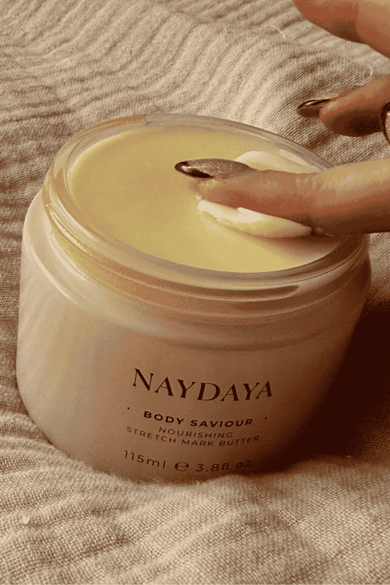 A close-up of a NAYDAYA Body Saviour Stretch Mark Butter jar with a hand applying the cream to skin, set against a textured beige linen background.