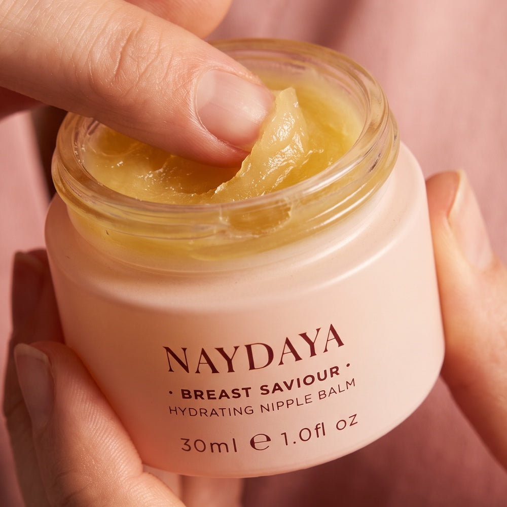 A close-up of a hand holding a jar of NAYDAYA Breast Saviour Hydrating Nipple Balm, with another hand lifting a piece of the yellow balm from the open jar.