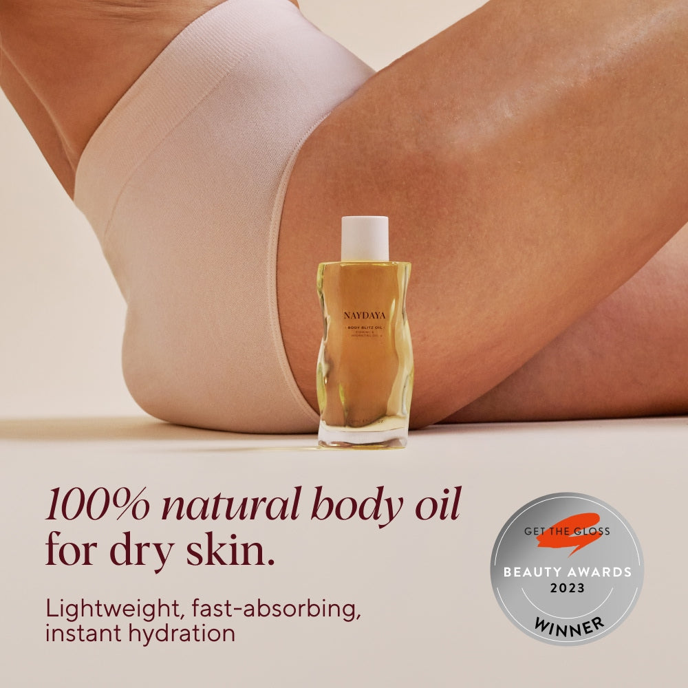 A bottle of Naydaya Natural Body Blitz Oil stands on a beige surface in front of a person's legs wearing light beige underwear. Text on the image reads '100% natural body oil for dry skin.