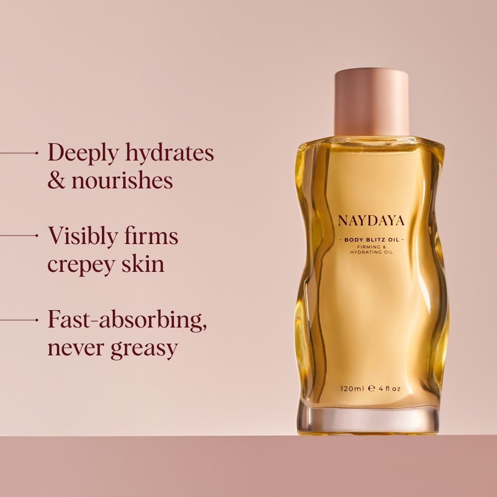 A bottle of NAYDAYA Body Blitz Oil shown on a soft beige background with marketing text on the left stating 'Deeply hydrates', 'visibly firms crepey skin' and 'fast absorbing'. The bottle is transparent and filled with golden-yellow oil, topped with a pale pink cap.