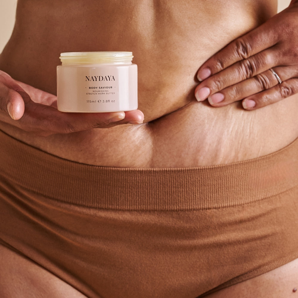 A close-up of a person's midsection holding a small cream-colored jar labeled NAYDAYA BODY SAVIOUR against their skin, with one hand holding the product and the other touching the skin.