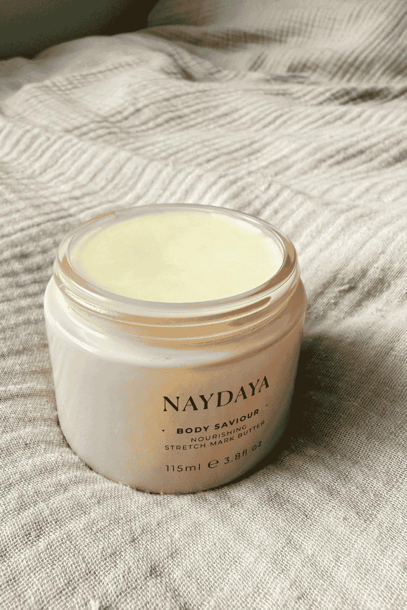 An open jar of NAYDAYA Body Saviour moisturising body butter, showing the pale yellow cream inside, set on a textured beige linen fabric background.