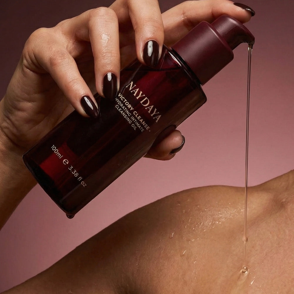 A hand with dark metallic nail polish holds a dark burgundy pump bottle of NAYDAYA Victory Cleanse Wash over a person's shoulder, with a thin stream of clear serum dripping onto the skin against a dusty rose background.
