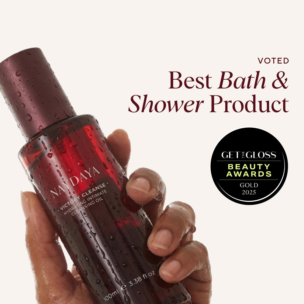 A hand holds a red Naydaya bath and shower oil bottle with water droplets on it against a light beige background, with promotional text reading 'Voted Best Bath & Shower Product' and a GetGloss Beauty Awards Gold 2025 badge.