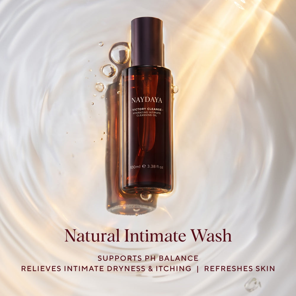 Naydaya Victory Cleanse intimate cleansing oil wash in a dark amber bottle with a black cap, centered over a glossy white liquid surface with ripples and bubbles. Text below reads 'Natural Intimate Wash' with supporting claims.